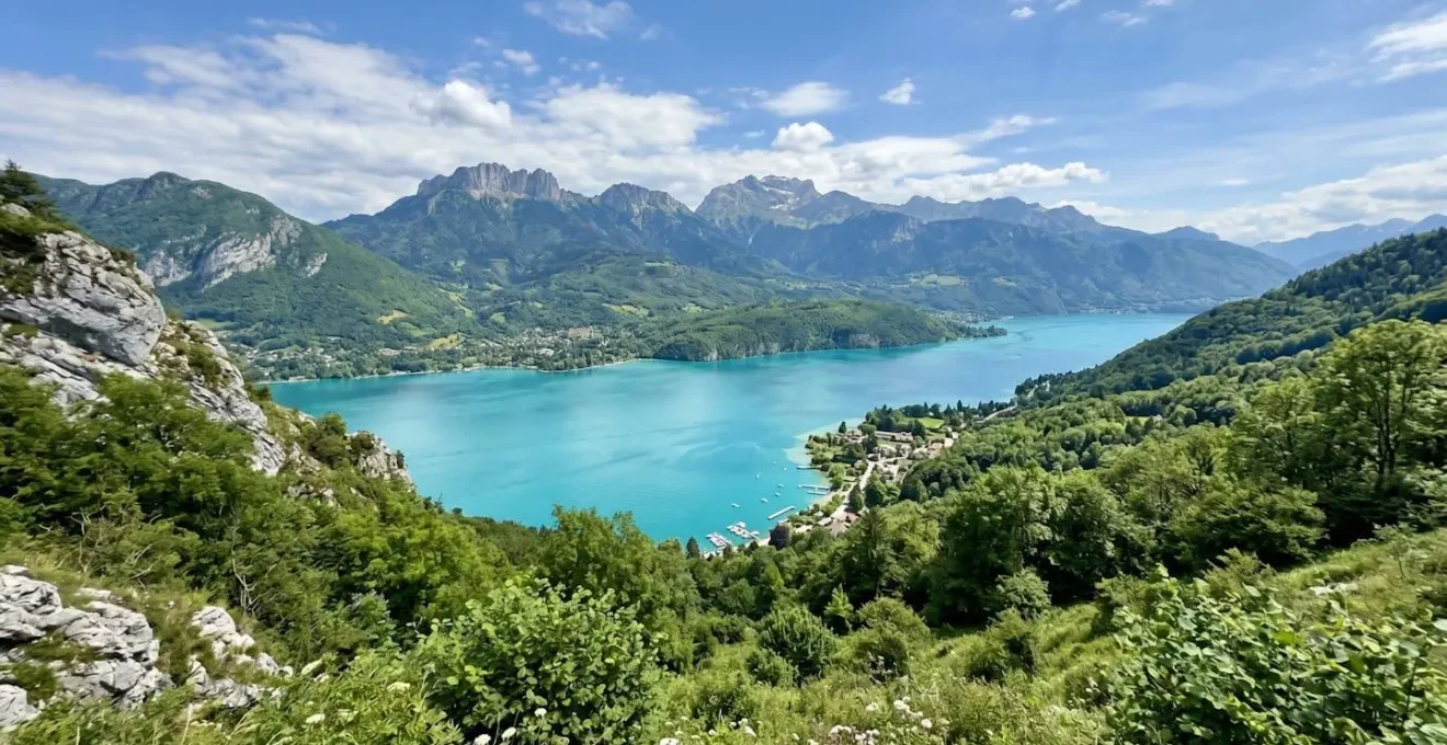 Vue large du lac d'Annecy depuis un point de vue élevé, eau turquoise au premier plan, montagnes enneigées en arrière-plan, végétation verte des rives et ciel bleu dégagé